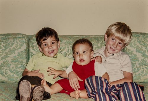 Three kids sitting on a couch, 2 young boys and a baby between them.